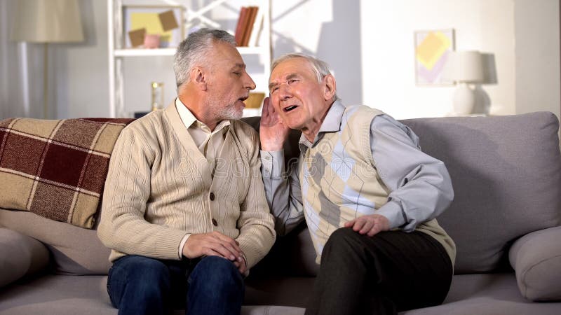 Old man talking to deaf friend sitting on sofa at home, hearing disease, problem