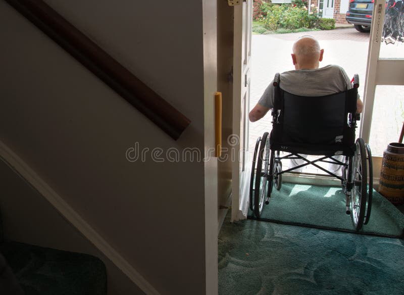 Old man in wheelchair looking out from the open doorway of his house.Hampshire,United Kingdom