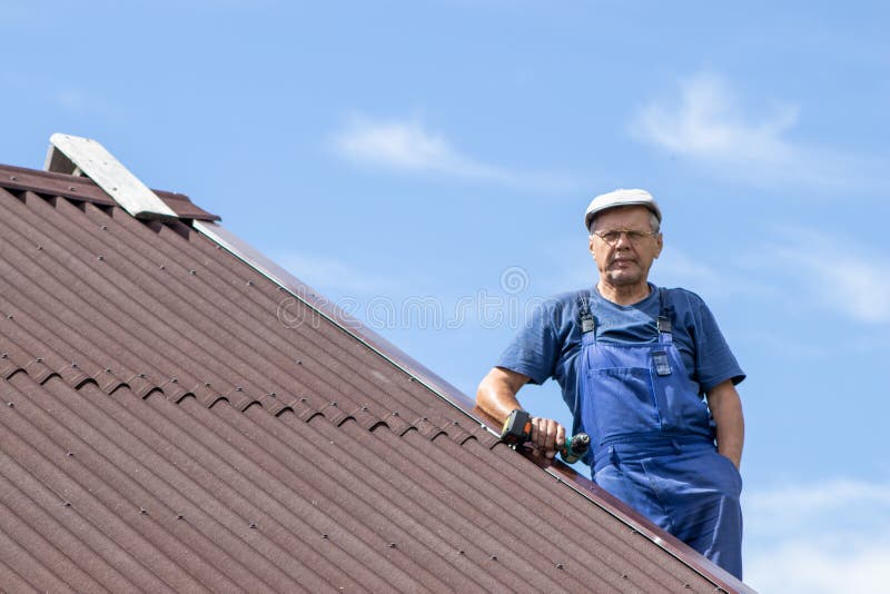 Old man working with electric screwdriver on a roof of a house with no safety harness, wearing work clothes, blue overall, dangero