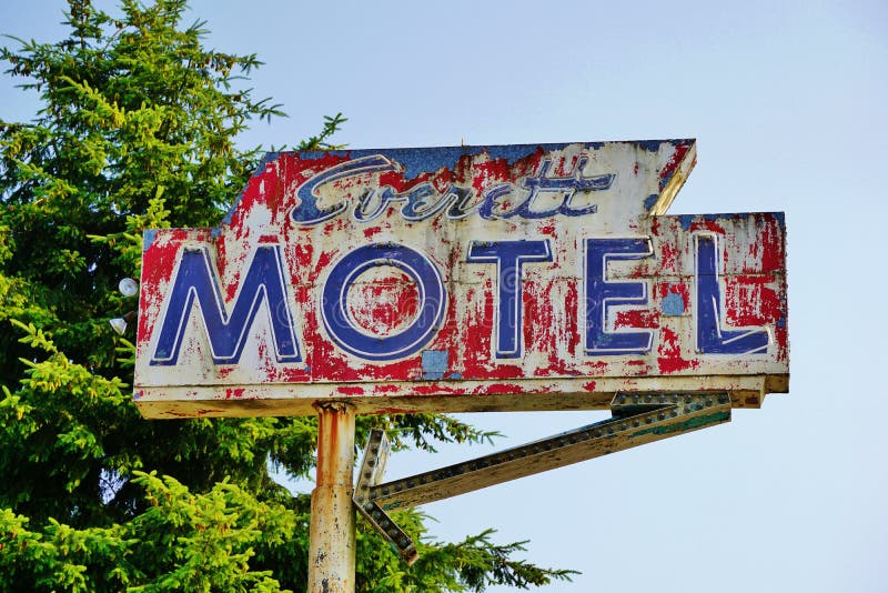 Outside an abandoned motel a neon and peeling painted red motel sign and directional arrow hang high up on a rusted metal pole with an evergreen tree and blue sky in the background. White neon letters over blue peeled painted lettering on a round metal three dimensional sign stands apart from the tree branches and clear blue sky in the background. The arrow is missing bulbs. Outside an abandoned motel a neon and peeling painted red motel sign and directional arrow hang high up on a rusted metal pole with an evergreen tree and blue sky in the background. White neon letters over blue peeled painted lettering on a round metal three dimensional sign stands apart from the tree branches and clear blue sky in the background. The arrow is missing bulbs.