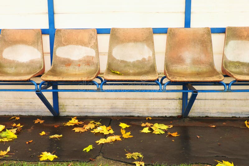 Old plastic seats on outdoor stadium players bench, chairs with worn paint below yellow roof. End of football seasson.