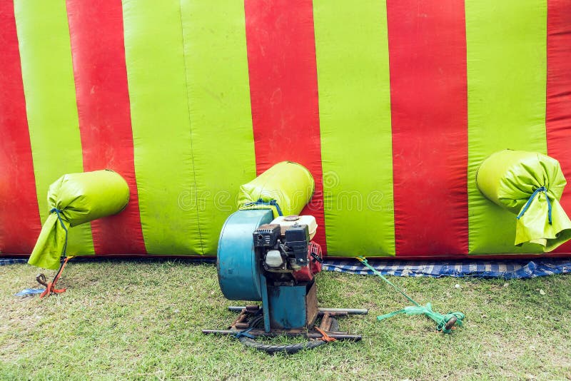 The old portable air blower pump fan install with green and red inflatable bouncer ball house castle or toy. Outdoor blue electric blower in the playground. Motor Air Blower on lawn in kid play land