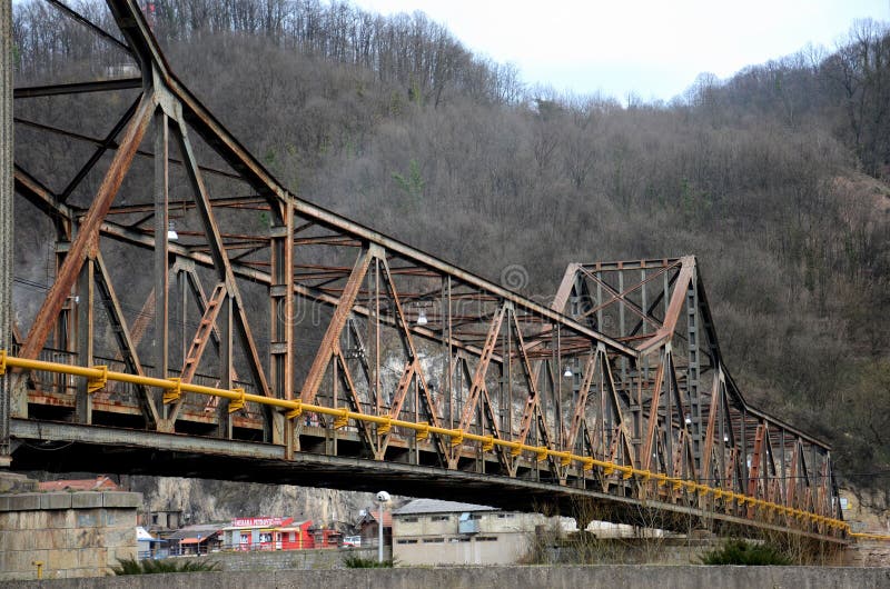 Bosnia Hercegovina - March 22, 2015: A weathered rusty old metal bridge across a river in Bosnia Hercegovina. In the background is a small town with some shops and wooded mountains. The bridge is lined with a freshly painted yellow pipe on its side. Bosnia Hercegovina - March 22, 2015: A weathered rusty old metal bridge across a river in Bosnia Hercegovina. In the background is a small town with some shops and wooded mountains. The bridge is lined with a freshly painted yellow pipe on its side.