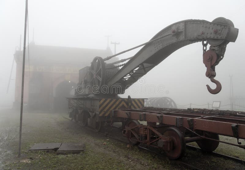 An old Train Crane still in use. A thick fog is over the Crane and a small car. A train workshop is on the back partially visible through the thick fog. City of Paranapiacaba, Sao Paulo state, Brazil.