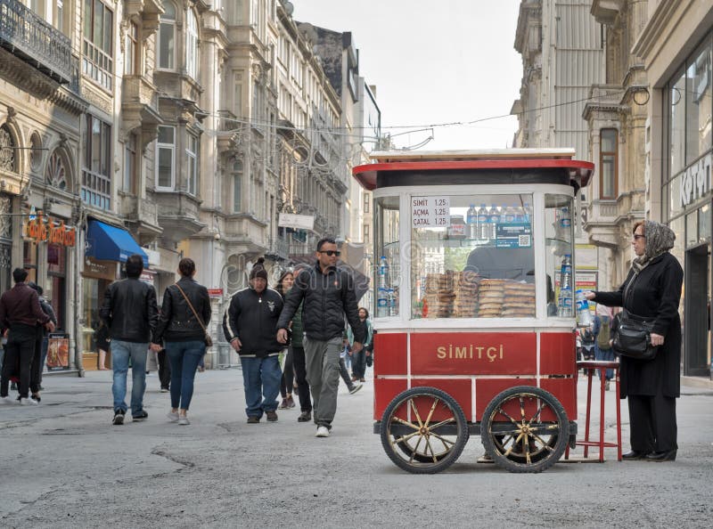 Istanbul, Turkey - April 24, 2017: Old Turkish lady buying fast food meal from a traditional Turkish Simit Turkish Bagel cart in Istiklal street and people walking at afternoon. Istanbul, Turkey - April 24, 2017: Old Turkish lady buying fast food meal from a traditional Turkish Simit Turkish Bagel cart in Istiklal street and people walking at afternoon