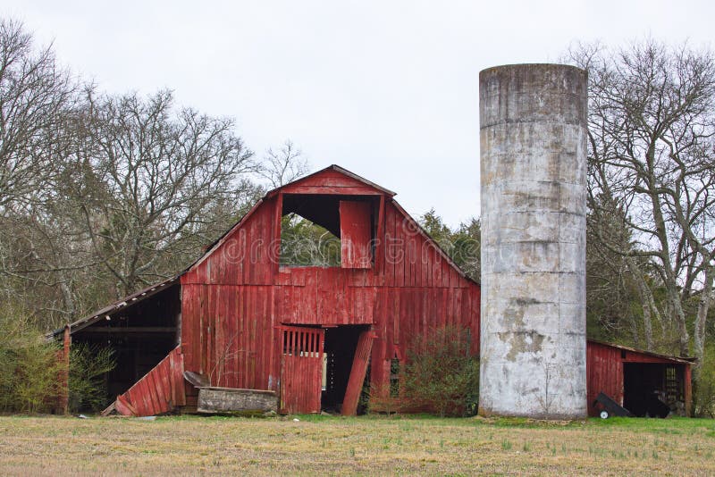 An old weathered red barn with a tin roof and a silo.  The barn has decorative front doors.  A concrete water trough is on the left side of the front of the barn. An old weathered red barn with a tin roof and a silo.  The barn has decorative front doors.  A concrete water trough is on the left side of the front of the barn