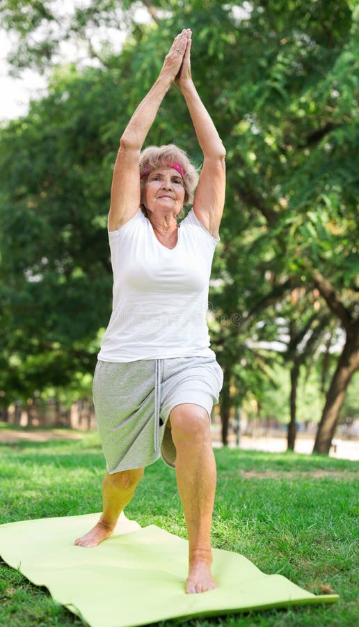 Old woman standing on mat in warrior I yoga pose in urban park. Old woman standing on mat in warrior I yoga pose in urban park