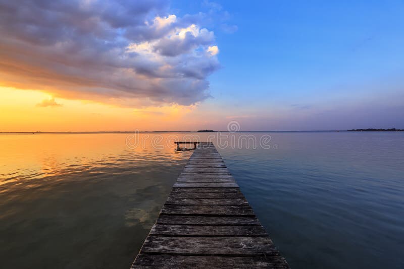 Old wooden jetty, pier reveals views of the beautiful lake, blue sky with cloud. Sunrise enlightens the horizon with orange warm
