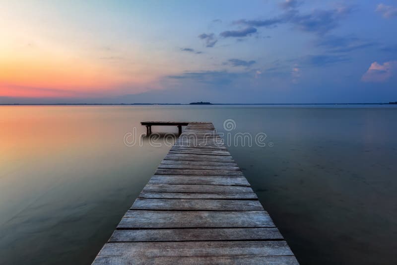 Old wooden jetty, pier reveals views of the beautiful lake, blue sky with cloud. Sunrise enlightens the horizon with orange warm