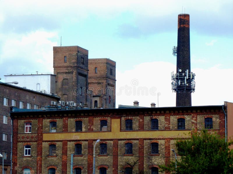 Old yellow, red and brown brick factory building exterior detail with arched windows, steel bar windows, tall industrial brick smoke stack and square brick towers in the background under pale blue and white sky. abstract view. Old yellow, red and brown brick factory building exterior detail with arched windows, steel bar windows, tall industrial brick smoke stack and square brick towers in the background under pale blue and white sky. abstract view.