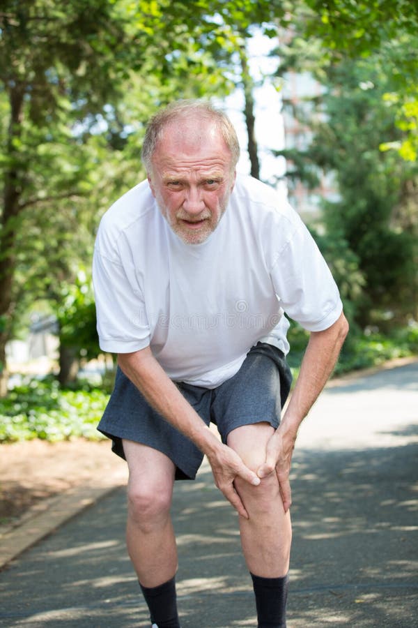 Closeup portrait, older man in white shirt, gray shorts, standing on paved road, in severe knee pain, isolated trees outside outdoors background. Closeup portrait, older man in white shirt, gray shorts, standing on paved road, in severe knee pain, isolated trees outside outdoors background.