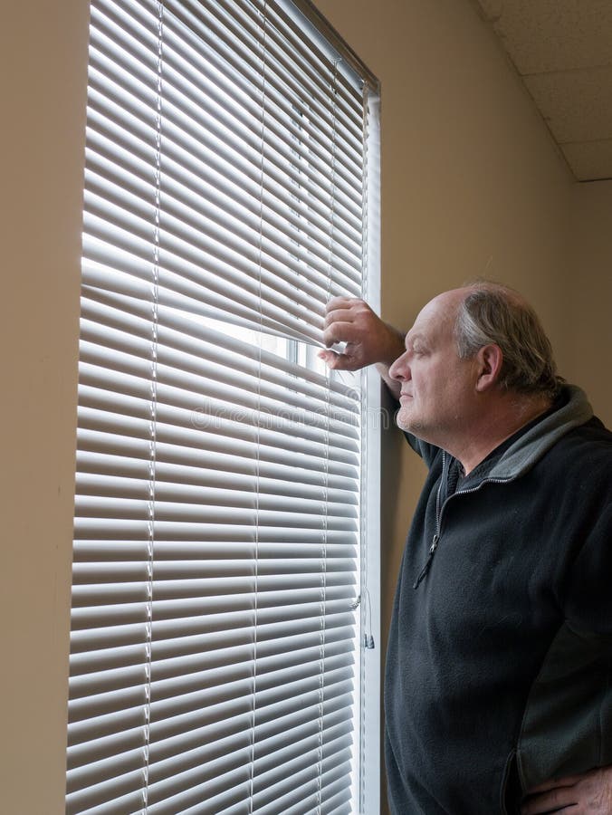 Older man looking out window blinds.