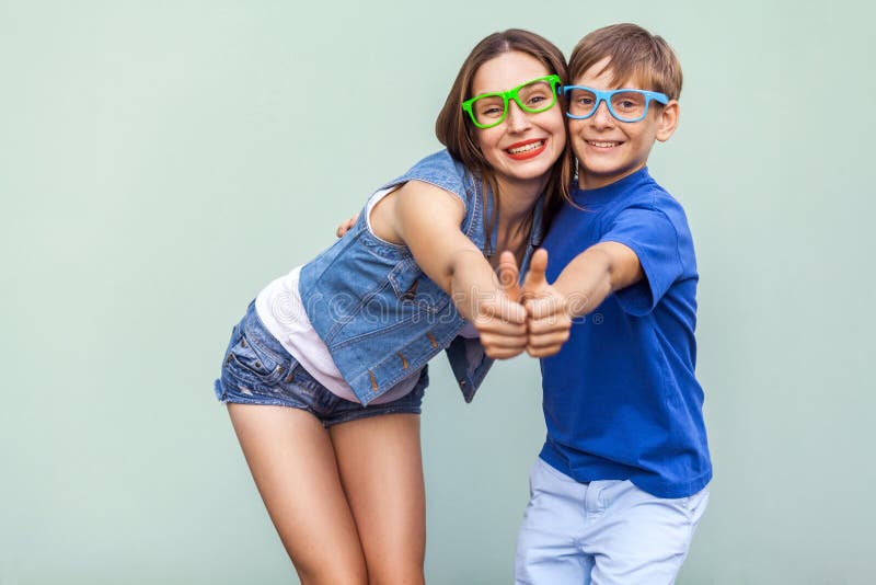 Older sister and her brother with freckles, posing over blue background together, looking at camera with toothy smile and thumbs