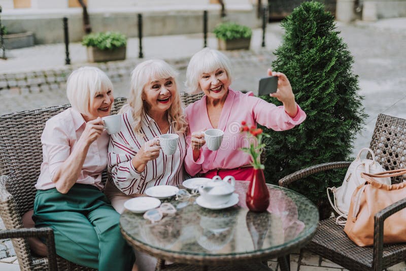 Stylish old ladies holding cups of tea and taking selfie on smartphone roya...