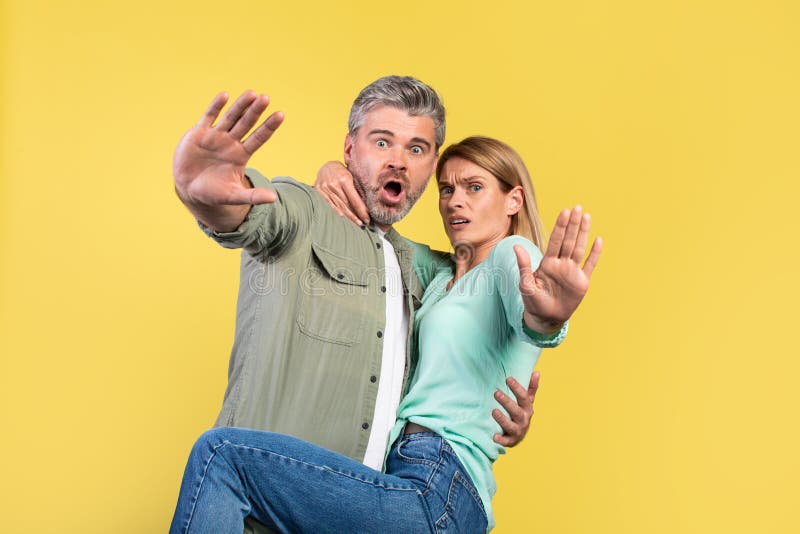 Omg. Portrait of scared middle aged spouses screaming and gesturing stop, woman jumping on husband, looking at camera over yellow studio wall. Human emotions, facial expression