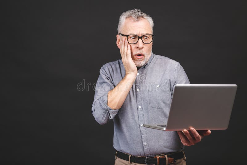 OMG! I`m shocked! Scared senior aged bearded handsome old man in eyeglasses holding laptop computer isolated against black background. OMG! I`m shocked! Scared senior aged bearded handsome old man in eyeglasses holding laptop computer isolated against black background