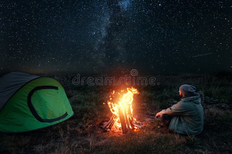 Omul de camping stă noaptea lângă foc pe fundalul cerului înstelat. conceptul de turism turistic în camping royalty free stock images