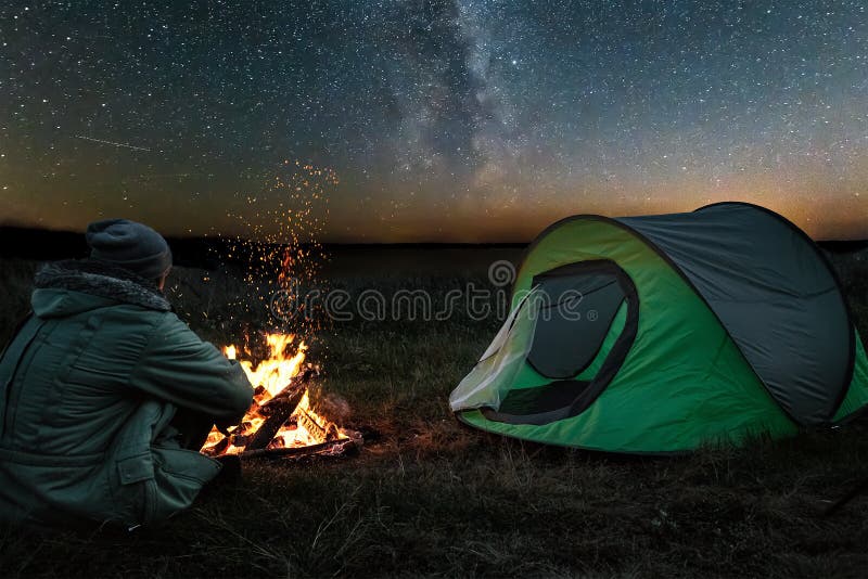Omul de camping stă noaptea lângă foc pe fundalul cerului înstelat. conceptul de turism turistic în camping stock photography