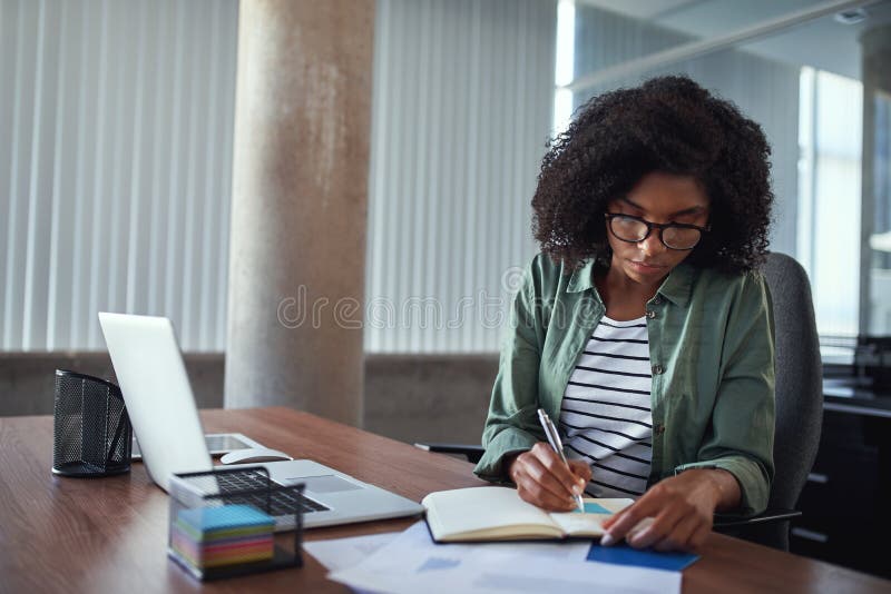 Close-up of an african young businesswoman writing schedule in diary over the wooden desk in the office. Close-up of an african young businesswoman writing schedule in diary over the wooden desk in the office