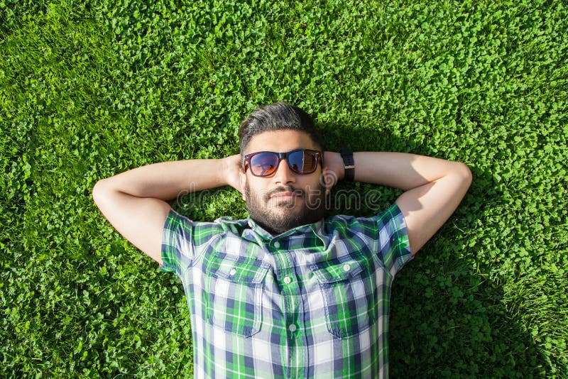 One fashion middle eastern man with beard, fashion hair style is resting on beautiful green grass day time.