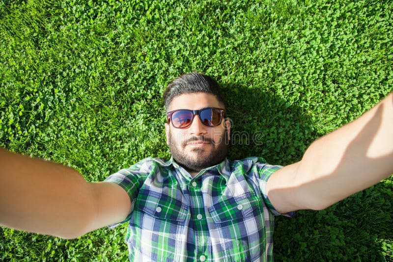 One young fashion middle eastern man with beard and fashion hair style is lying on a grass in a park taking selfie. stock images One young fashion middle eastern man with beard and fashion hair style is lying on a grass in a park taking selfie. stock images