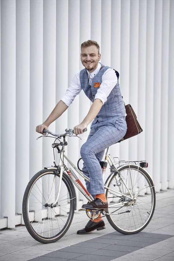 One young man, 20-29 years old, wearing hipster suit, smart casual, posing on old city bike. white modern wall building behind.