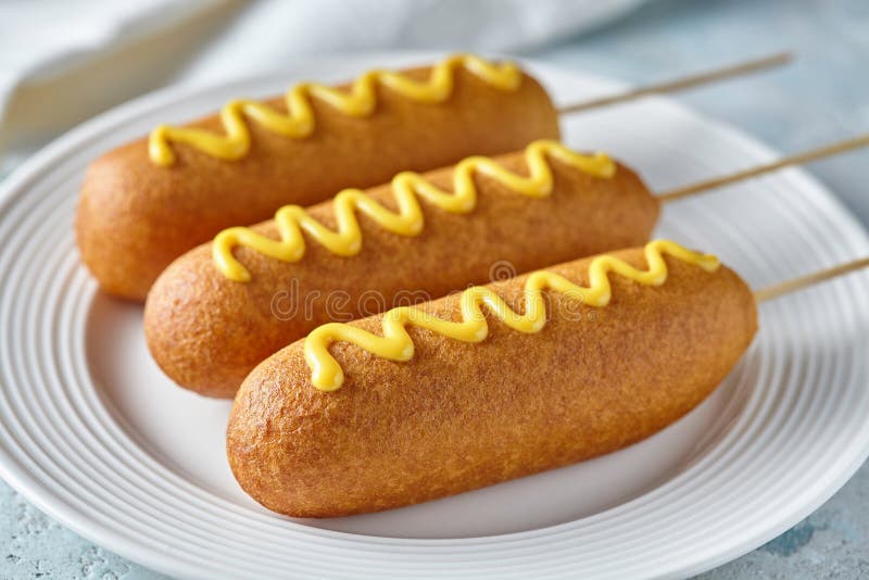 Corn dog traditional American corndog street junk food fried hotdog meat wiener with mustard on top snack treat in a layer of cornmeal batter on stick unhealthy eating on rustic table. Corn dog traditional American corndog street junk food fried hotdog meat wiener with mustard on top snack treat in a layer of cornmeal batter on stick unhealthy eating on rustic table.