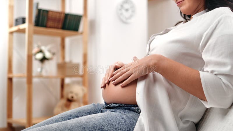 Onherkenbare glimlachende brunette vrouw die de zwangere buik aanraakt die op een bank zit in een witte lichtkamer