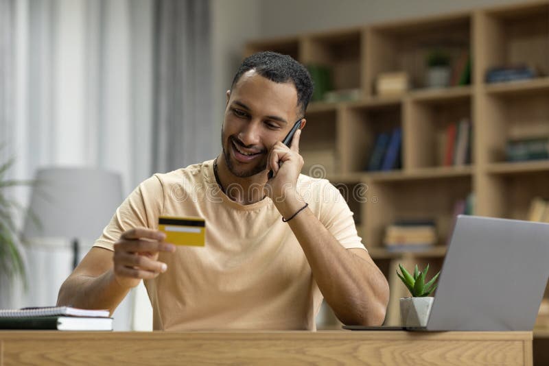 Online shopping. Happy arab man using credit card and laptop, talking on phone, calling to bank for payment verification royalty free stock images