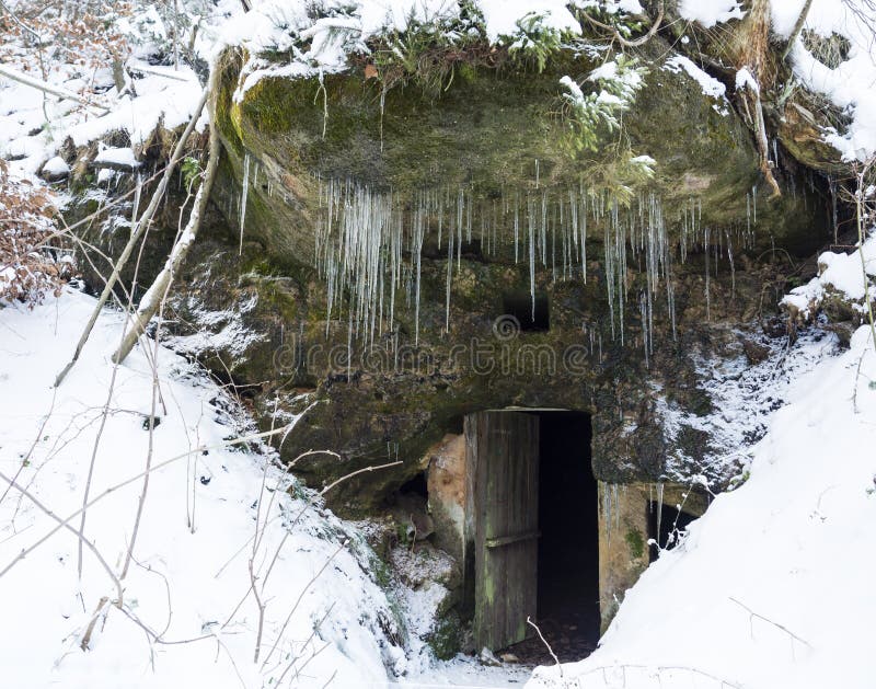 Open donkere kelderdeur in het hol van de zandsteenrots met mos en korstmossen en lange hangende ijskegels, witte sneeuwachtergro royalty-vrije stock foto Open donkere kelderdeur in het hol van de zandsteenrots met mos en korstmossen en lange hangende ijskegels, witte sneeuwachtergro royalty-vrije stock foto