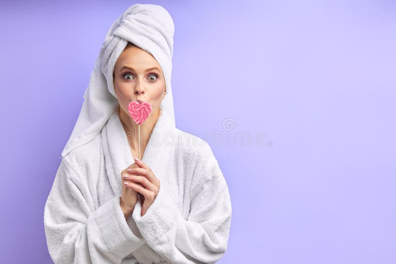 Open-eyed Young woman in towel and bathrobe with lollipop isolated over purple background