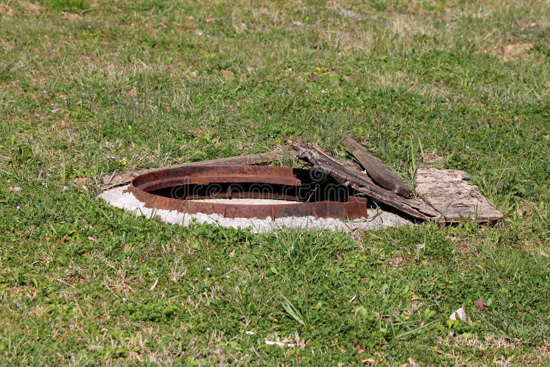 Open manhole with rusted frame and broken wooden boards surrounded with uncut grass stock image