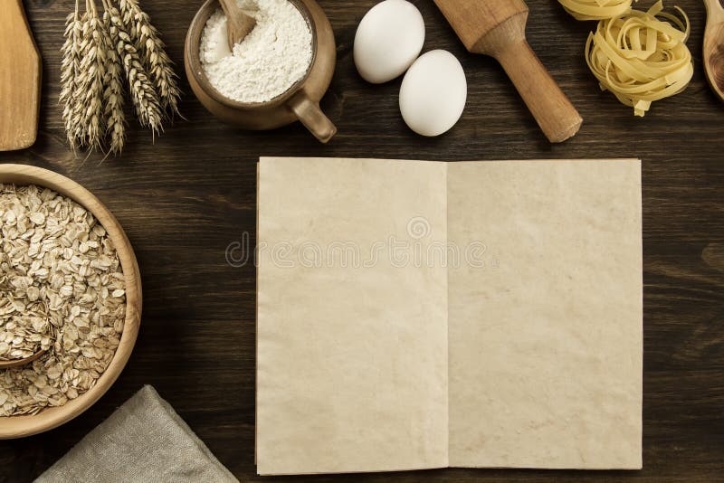 Open old vintage book on the aged wooden background. Kitchen utensils, ears of wheat, flour in a pot.