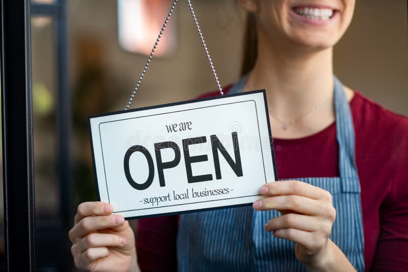 Small business owner smiling while turning the sign for the reopening of the place after the quarantine due to covid-19. Happy businesswoman standing at her restaurant or coffee shop gate with open signboard. Close up of woman’s hands holding sign now we are open support local business. Small business owner smiling while turning the sign for the reopening of the place after the quarantine due to covid-19. Happy businesswoman standing at her restaurant or coffee shop gate with open signboard. Close up of woman’s hands holding sign now we are open support local business