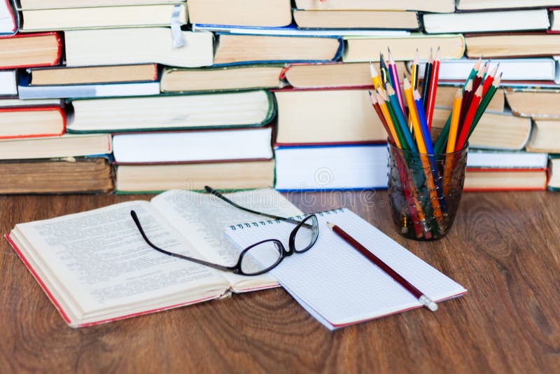 Open textbook, notebook, glasses, pencils in holder, stack of old book on wooden table, education concept background, many books