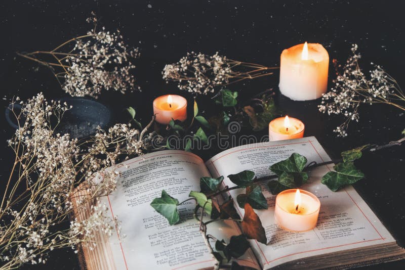 Open vintage poetry book on a black table surface with white lit burning candles and dried flowers. Dark, romantic and cozy feel with a branch of ivy on top of