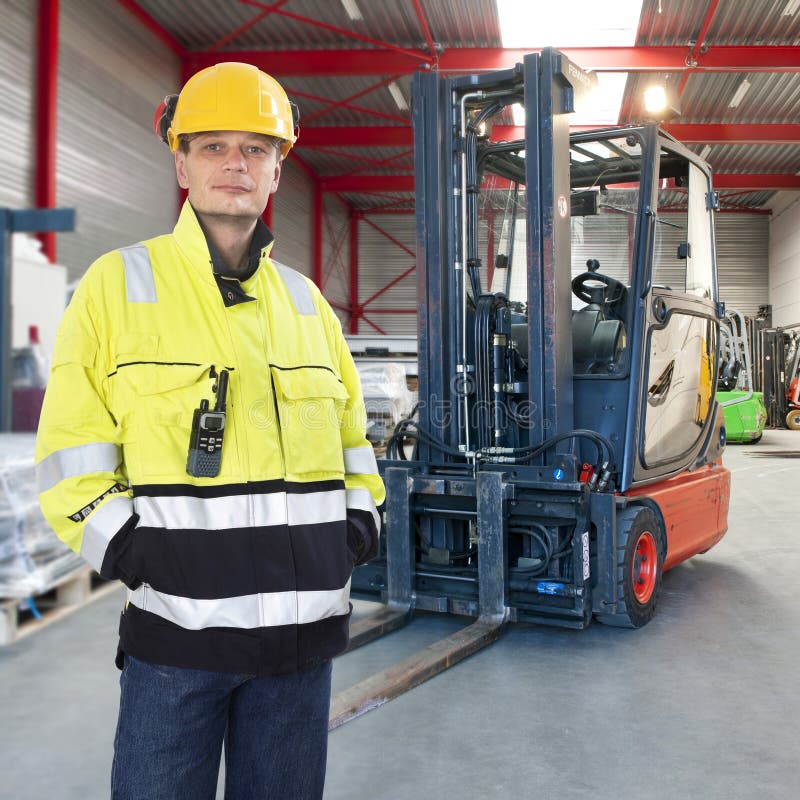 Man, waring safety clothes, including a hard hat, standing proudly in front of his forklift truck in a warehouse. Man, waring safety clothes, including a hard hat, standing proudly in front of his forklift truck in a warehouse