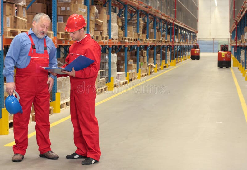 Standing workers in uniforms with hardhats, reading invoice in factory warehouse. one is older ,one is young. Standing workers in uniforms with hardhats, reading invoice in factory warehouse. one is older ,one is young