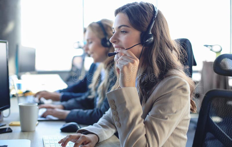 Operator de asistență pentru clienți femei cu căști și zâmbind cu colegii în fundal royalty free stock image