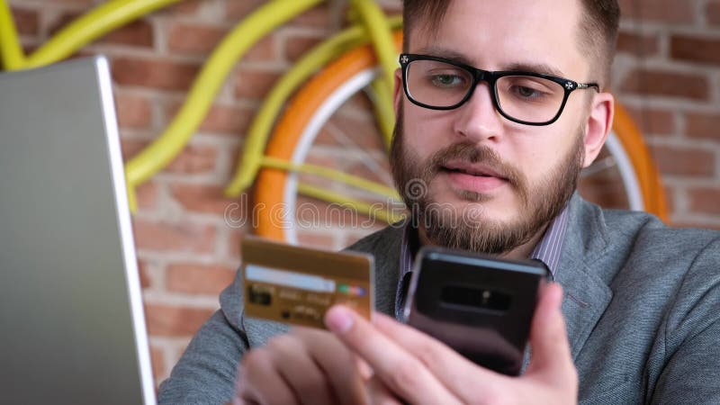 Operação bancária em linha com telefone esperto Um homem novo moderno elegante com uma barba usa um cartão de crédito bancário pa