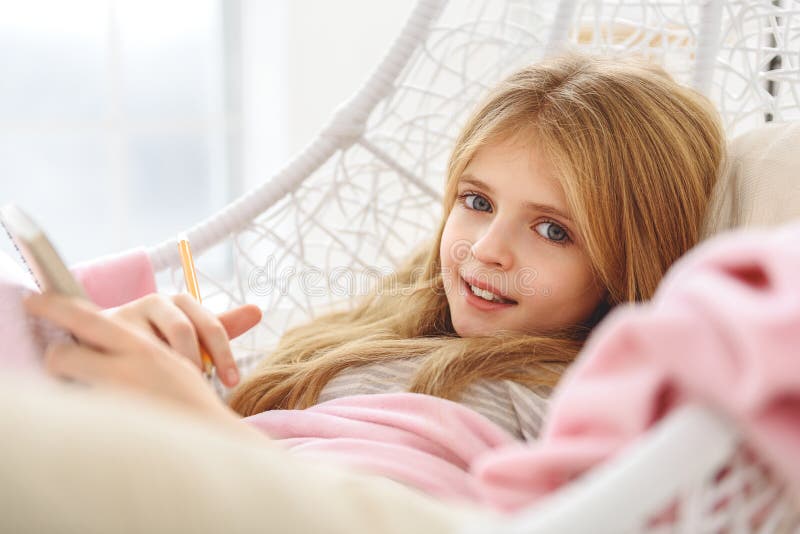 Happy girl is keeping a diary with excitement. She is holding notebook with pencil and smiling. Kid is lying on hammock. Happy girl is keeping a diary with excitement. She is holding notebook with pencil and smiling. Kid is lying on hammock