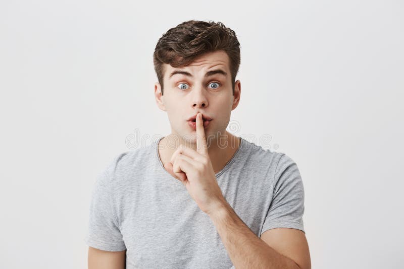 Excited surprised caucasian guy with stylish hairstyle casually dressed showing silence sign asking to be quiet. Indoor portrait of handsome attractive male with bugged eyes holding his finger on lips. Excited surprised caucasian guy with stylish hairstyle casually dressed showing silence sign asking to be quiet. Indoor portrait of handsome attractive male with bugged eyes holding his finger on lips.