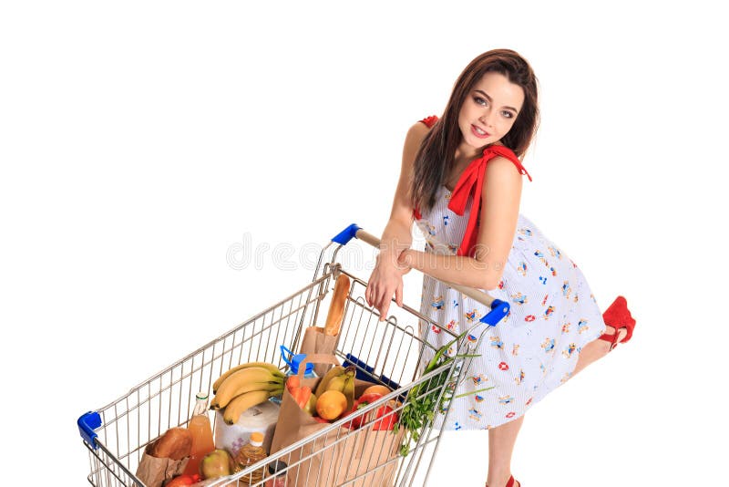 Opinião de ângulo alto a menina que sorri na câmera ao empurrar um carrinho de compras completamente com os mantimentos isolados foto de stock Opinião de ângulo alto a menina que sorri na câmera ao empurrar um carrinho de compras completamente com os mantimentos isolados foto de stock