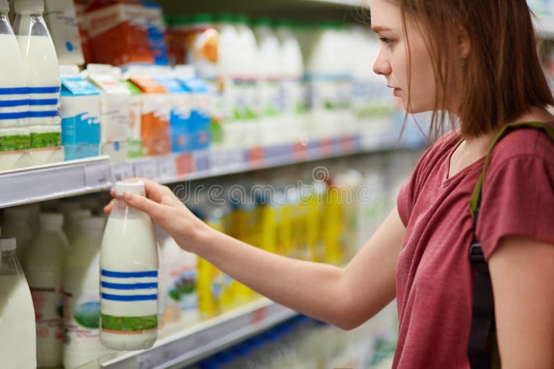 A opinião horizontal a fêmea nova bonita séria escolhe produtos de leite no deparment da leiteria do supermercado, vestido no shi fotografia de stock royalty free A opinião horizontal a fêmea nova bonita séria escolhe produtos de leite no deparment da leiteria do supermercado, vestido no shi fotografia de stock royalty free