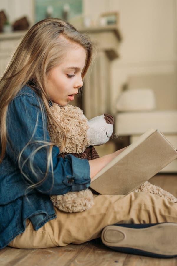 Opinião lateral a menina com o livro de leitura do urso de peluche imagem de stock royalty free Opinião lateral a menina com o livro de leitura do urso de peluche imagem de stock royalty free