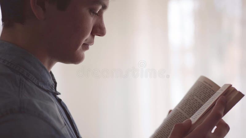 Opinião lateral o homem novo que lê livro interessante na sala na frente do grande fim da janela acima Lazer em casa Conceito