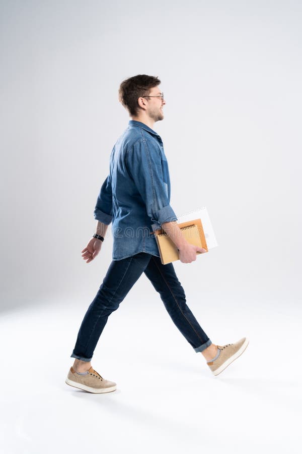 Opinião lateral um homem ocasional novo de sorriso que anda, estudante com notas do ANG do livro no fundo branco fotografia de stock Opinião lateral um homem ocasional novo de sorriso que anda, estudante com notas do ANG do livro no fundo branco fotografia de stock