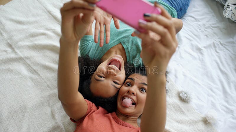 Opinião superior as amigas engraçadas bonitas da raça misturada que fazem o retrato do selfie na cama no quarto em casa imagens de stock Opinião superior as amigas engraçadas bonitas da raça misturada que fazem o retrato do selfie na cama no quarto em casa imagens de stock