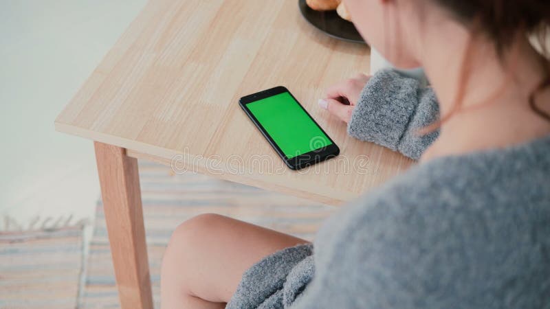 Opinião traseira a mulher que senta-se na tabela na cozinha em casa A menina moreno usa o smartphone, tela verde imagens de stock royalty free Opinião traseira a mulher que senta-se na tabela na cozinha em casa A menina moreno usa o smartphone, tela verde imagens de stock royalty free
