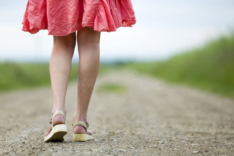 Opinión trasera la mujer joven en piernas rojas del vestido que camina por el camino de tierra el día de verano en fondo soleado foto de archivo Opinión trasera la mujer joven en piernas rojas del vestido que camina por el camino de tierra el día de verano en fondo soleado foto de archivo